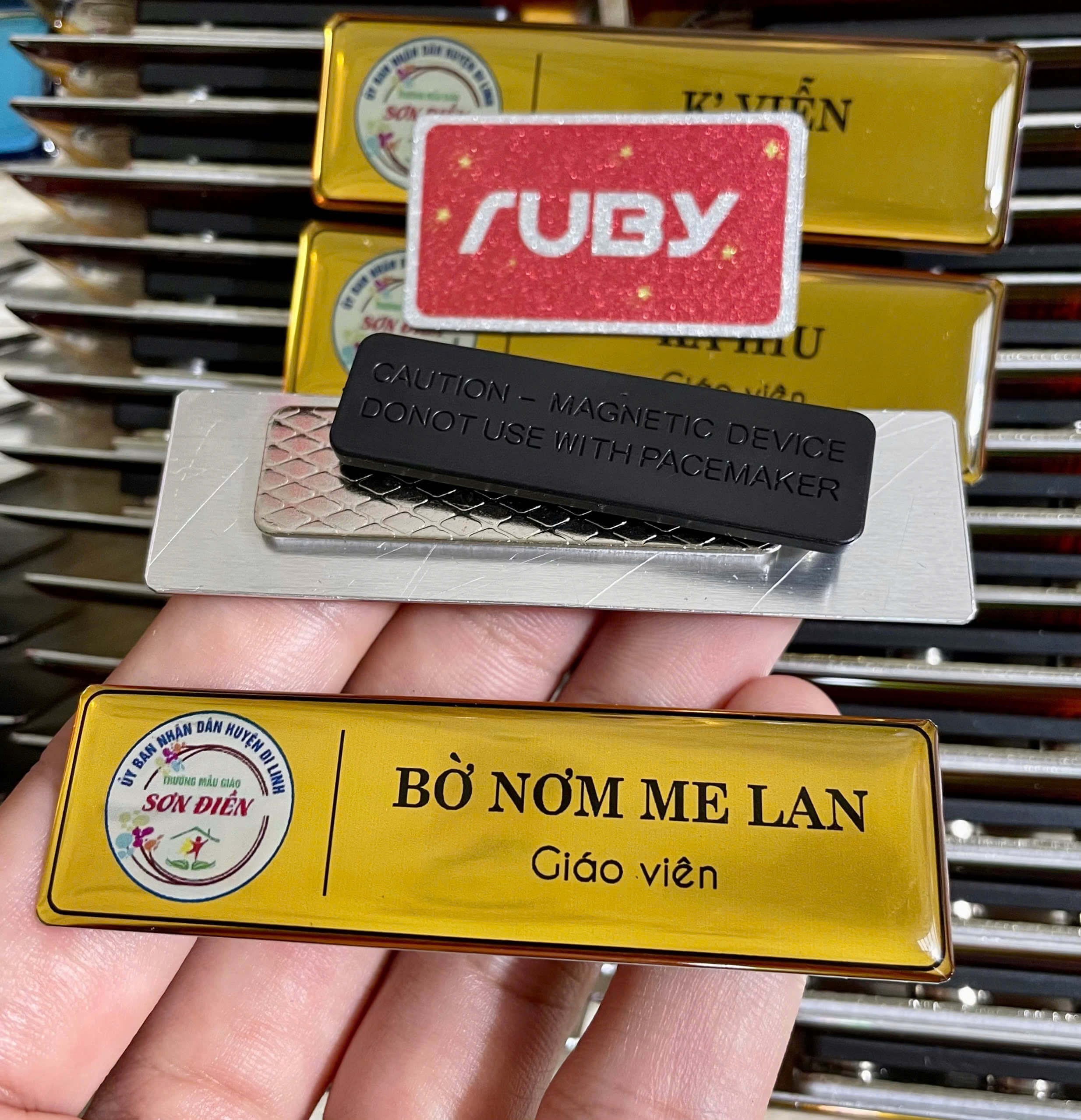 Bảng tên giáo viên nam châm bằng nhôm nền vàng của Trường Mẫu giáo Sơn Điền, in tên “Bờ Nơm Me Lan”; mặt sau nam châm 3 nút, không thủng áo.