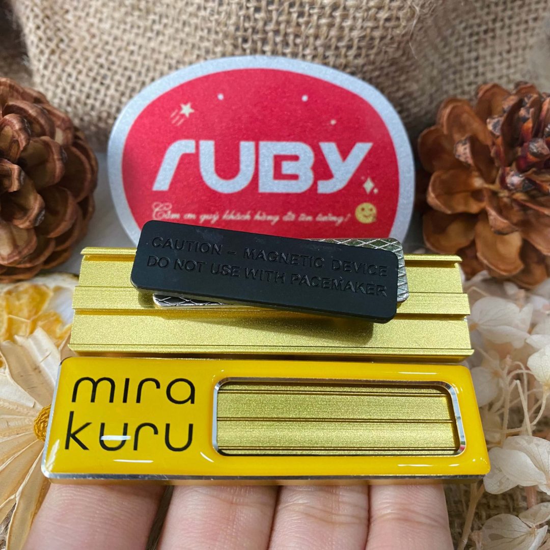 Bảng tên Mira Kuru màu vàng, có logo chữ đen, mặt sau gắn nam châm hít áo