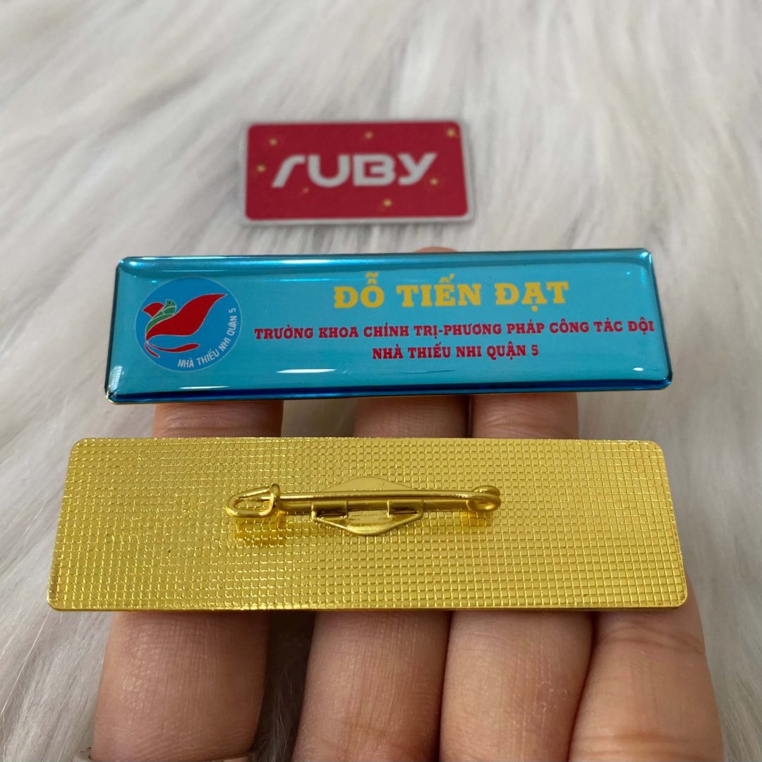 Bảng tên Nhà Thiếu Nhi Quận 5 màu xanh ngọc, in logo chim bồ câu, tên Đỗ Tiến Đạt – Trưởng khoa chính trị, mặt sau ghim cài áo vàng.