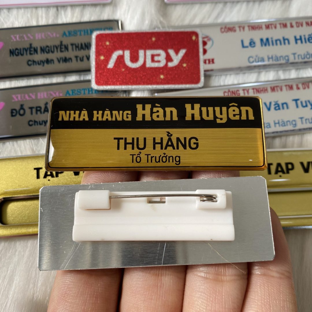 Bảng tên nhân viên nhà hàng Hàn Huyên màu vàng ánh kim, in logo và chức vụ, mặt sau ghim cài áo chắc chắn.