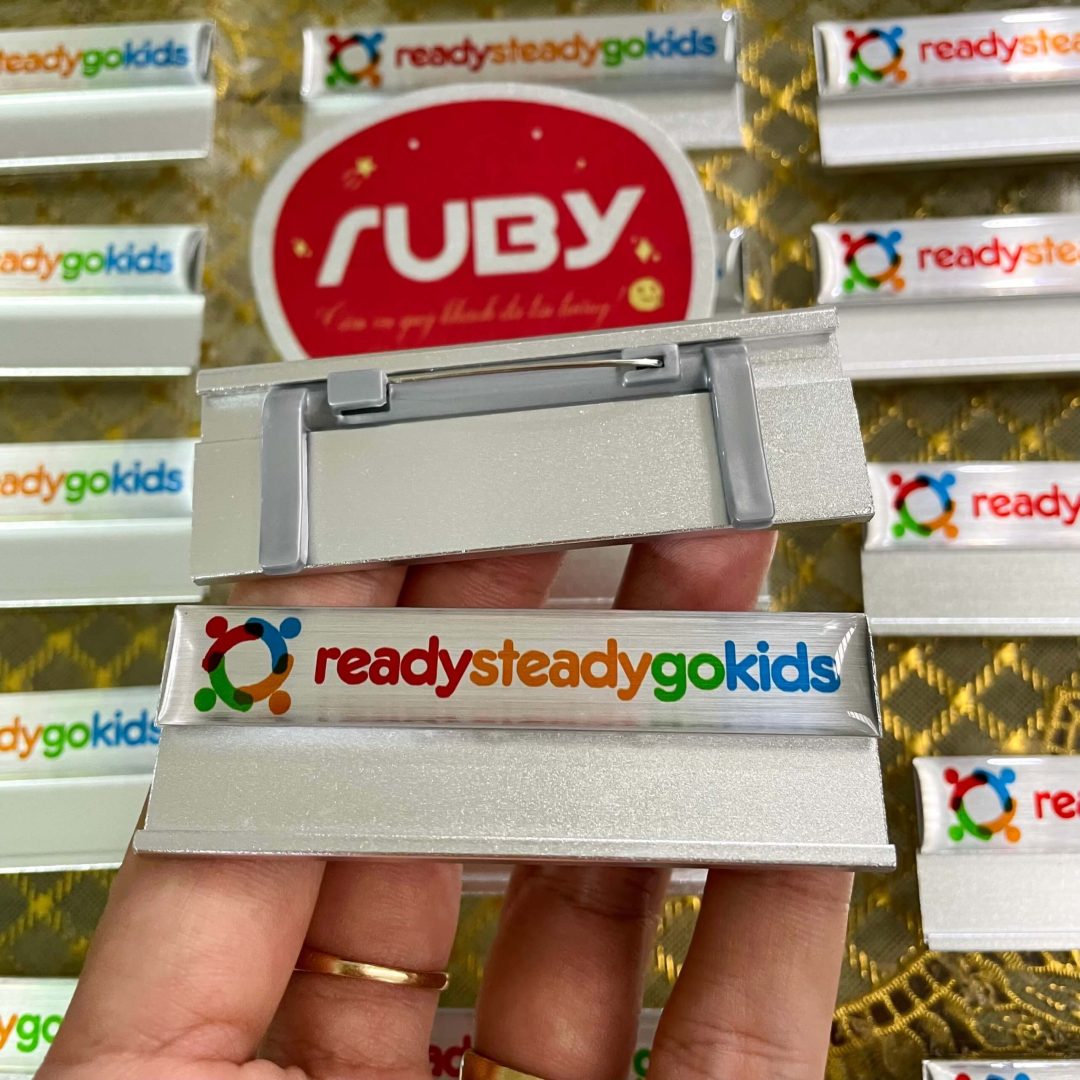 Bảng tên Ready Steady Go Kids, nền bạc, in logo màu tươi sáng, cài gài phía sau.