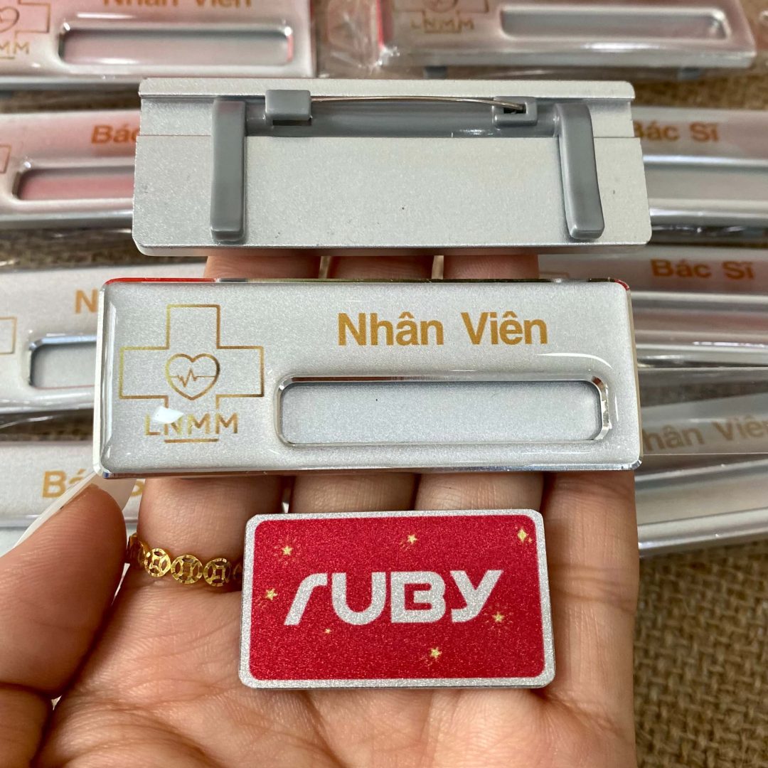 Bảng tên nhân viên LNMM, nền bạc, in logo chữ thập vàng, gài nhựa phía sau.