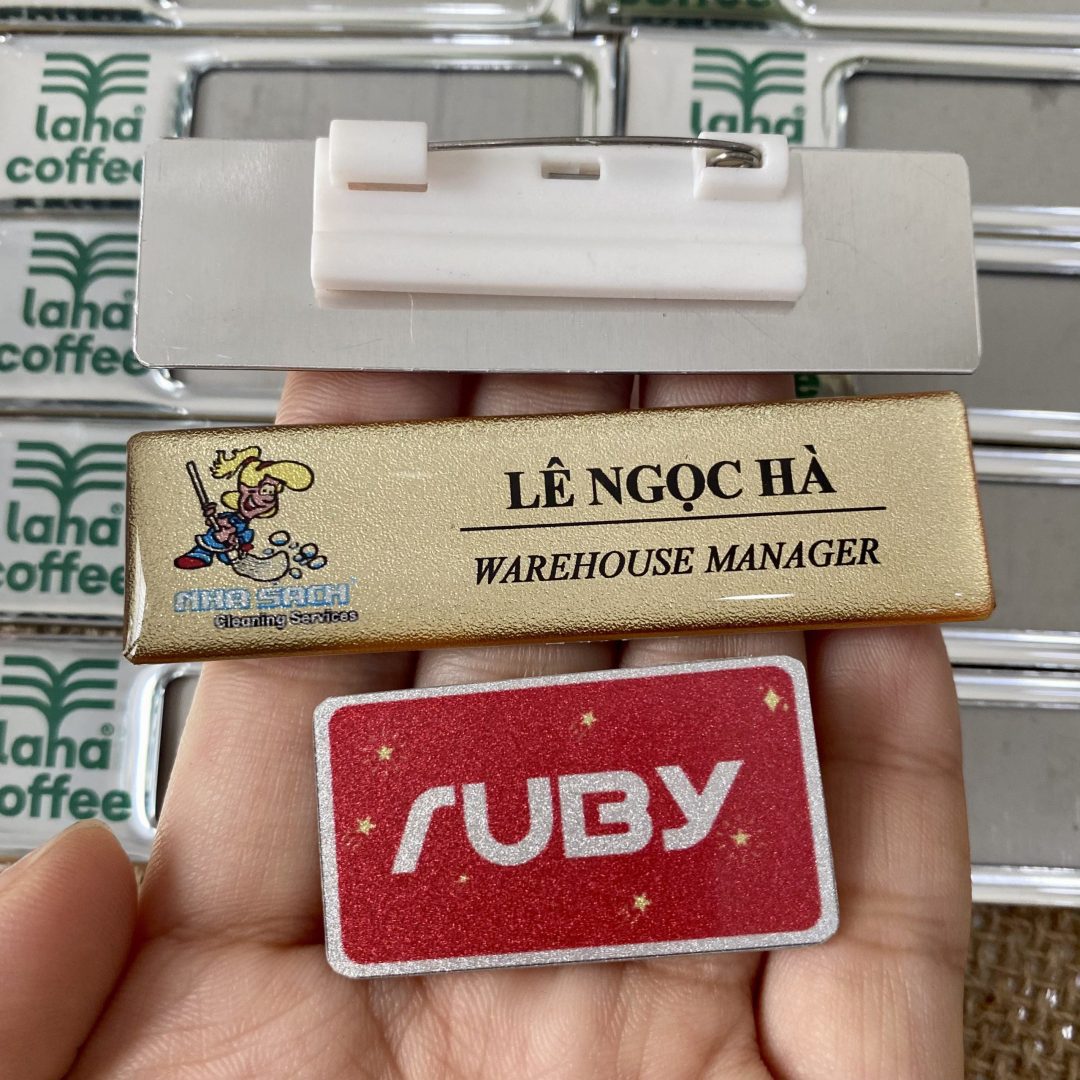 Bảng tên nhân viên công ty màu vàng, in logo hình nhân vật cầm chổi, tên Lê Ngọc Hà – Warehouse Manager, ghim cài áo.