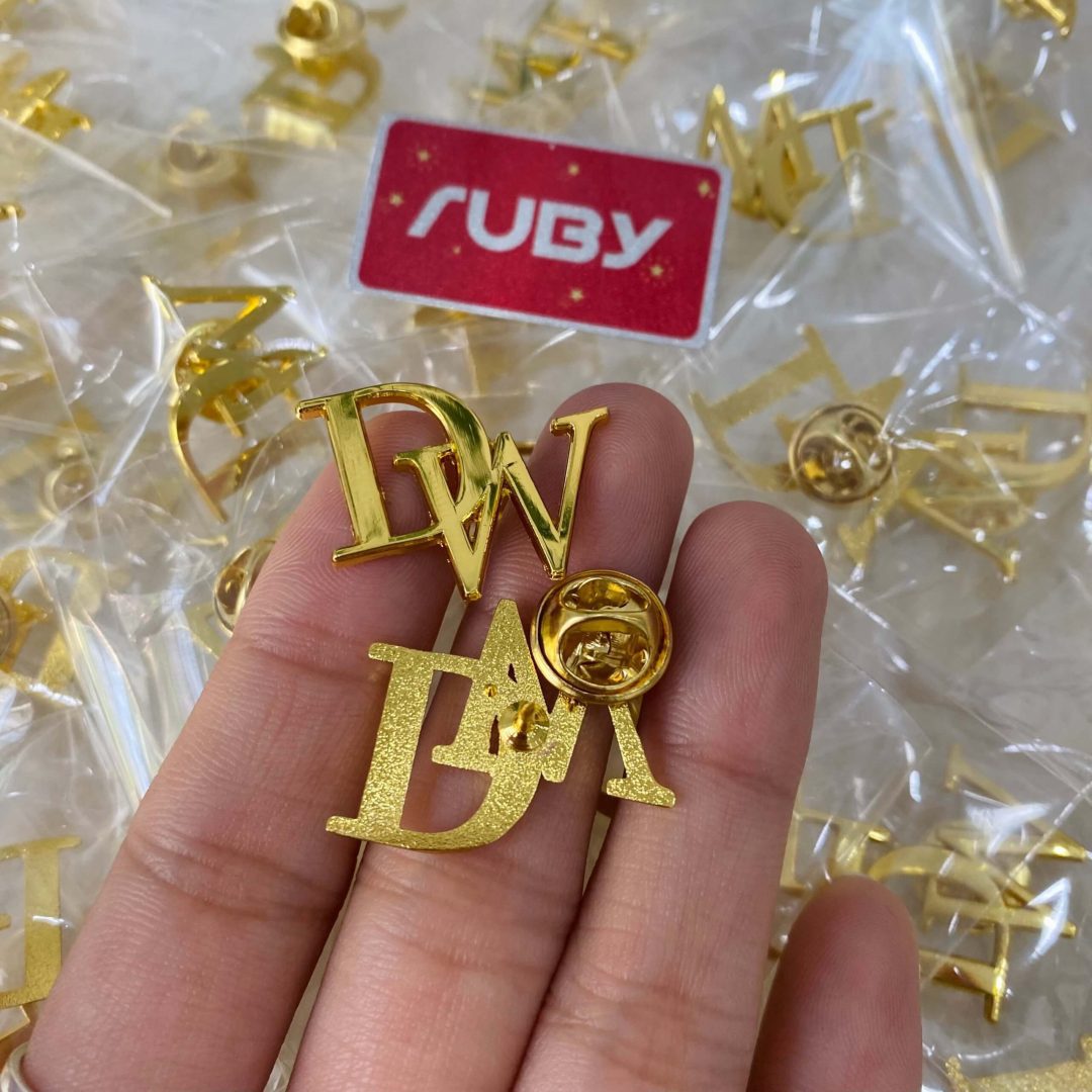 Huy hiệu chữ DW kim loại mạ vàng, chốt gài tròn, bề mặt sáng bóng và sang trọng